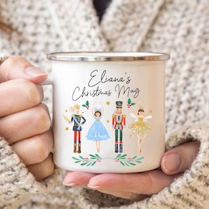 May include: White enamel mug with a festive Nutcracker theme. It features a ballerina, a nutcracker, a mouse king, and a sugar plum fairy. The mug has the text "Eliana's Christmas Mug" printed on it.