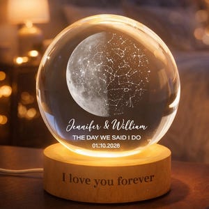 Puede incluir: Una bola de cristal brillante con un dise&ntilde;o de luna y constelaciones, sobre una base de madera. La base est&aacute; grabada con "I love you forever". La bola de cristal tiene los nombres "Jennifer & William" y la fecha "01.10.2026".