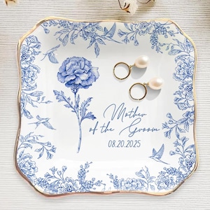 May include: A white and blue floral ceramic trinket dish with gold trim. The dish features a blue flower and the text "Mother of the Groom" with a date. Two gold earrings with pearls rest on the dish.