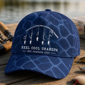 Op de afbeelding: Een marineblauwe baseballcap met een wit hengelontwerp en de tekst "REEL COOL GRANDPA". De cap heeft een netachtig patroon en de namen van familieleden. De tekst "EST. GRANDPA 2020" is ook opgenomen.