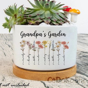 May include: White ceramic planter with the text "Grandpa's Garden" and names below illustrated flowers. The planter holds a succulent plant and decorative mushrooms. The planter sits on a round wooden base.