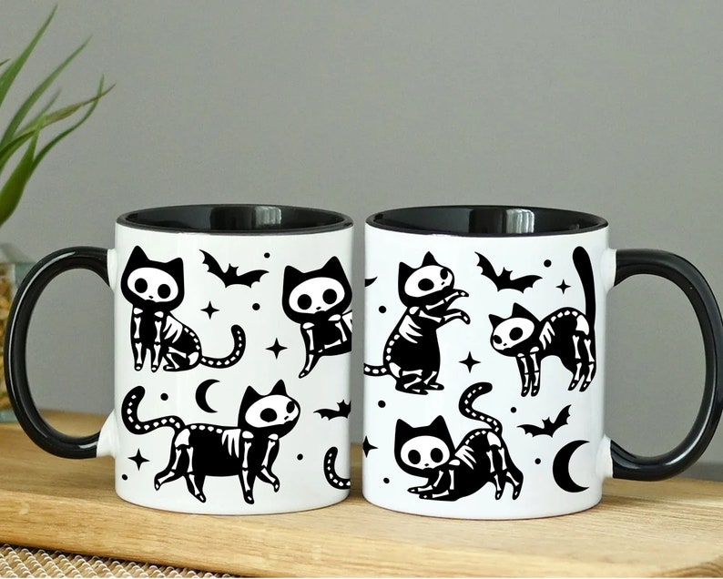 Pu&ograve; includere: Due tazze in ceramica bianca e nera con un bordo nero. Le tazze presentano un motivo ripetuto di gatti neri con scheletri bianchi e pipistrelli. Le tazze sono su una superficie di legno.