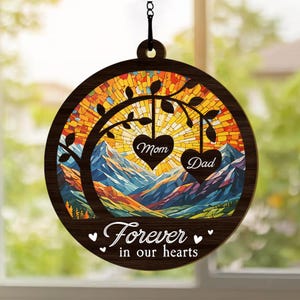 May include: A wooden suncatcher with a stained glass design of a tree with mountains in the background. The suncatcher has two hearts hanging from the branches, one that says "Mom" and one that says "Dad". The suncatcher also has the text "Forever in our hearts" at the bottom.