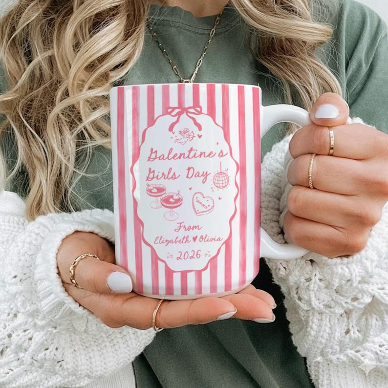 May include: White ceramic mug with pink vertical stripes. The mug features a pink oval design with the text "Galentine's Girls Day" and illustrations of cocktails and a heart-shaped cookie. The text "From Elizabeth & Olivia 2026" is also on the mug.