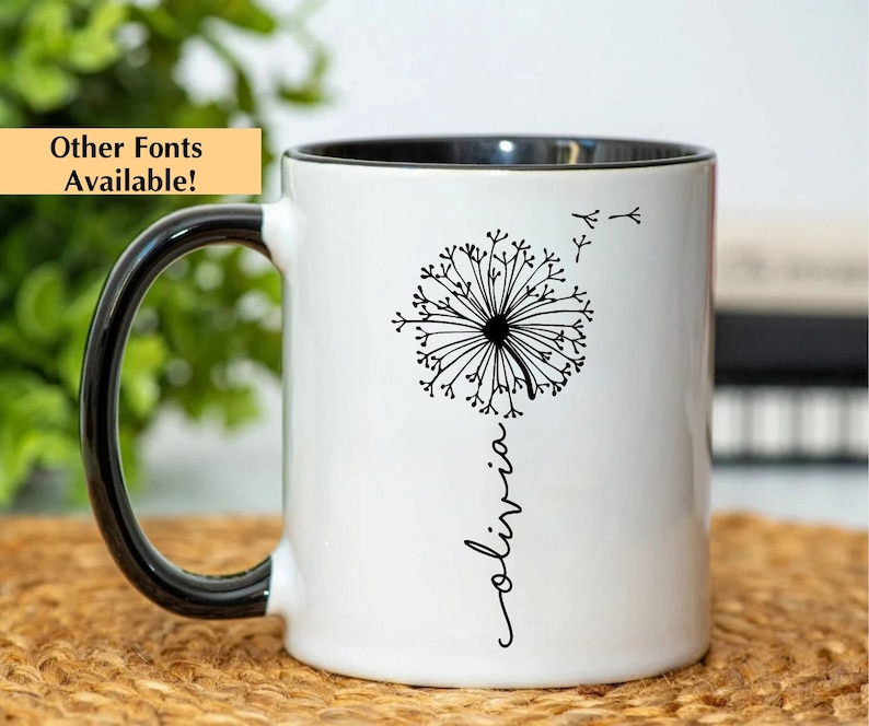 May include: White ceramic mug with a black rim and handle. The mug features a black and white illustration of a dandelion with the word "olivia" written vertically below it.