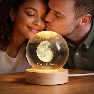 Puede incluir: Una esfera de cristal brillante con una imagen de la luna y el texto "OUR MOON". La esfera est&aacute; personalizada con los nombres "Sophia & Thomas" y la fecha "09.05.2023". Se encuentra sobre una base de madera, iluminada desde dentro.