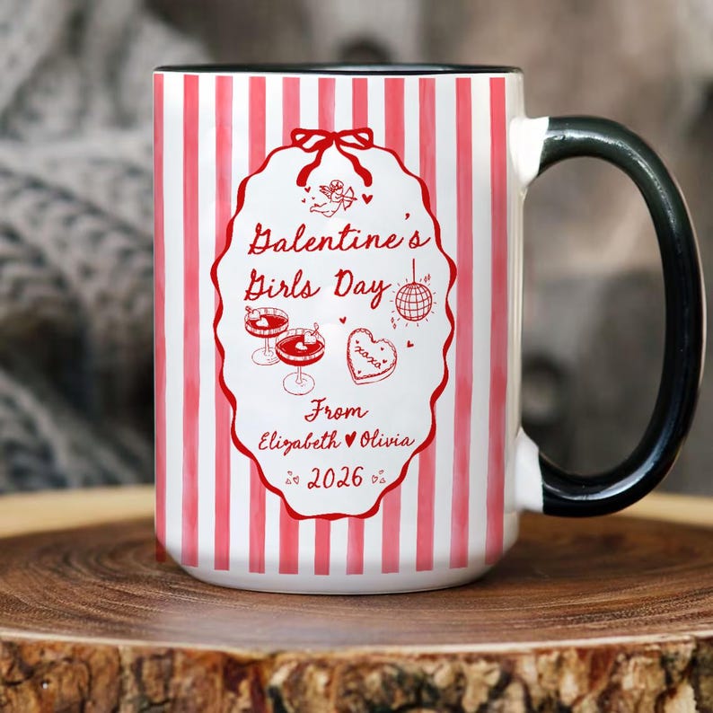 May include: A white ceramic mug with a black handle and rim, featuring red and white vertical stripes. The mug has a decorative oval with the text "Galentine's Girls Day" and other text and illustrations. The mug is a Valentine's Day gift.