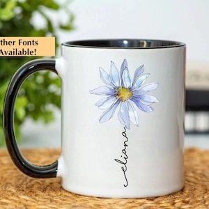Puede incluir: Taza de cer&aacute;mica blanca con asa y borde negros. La taza presenta una ilustraci&oacute;n de acuarela de una margarita azul con un centro amarillo y el nombre "eliana" escrito en cursiva debajo de la flor.