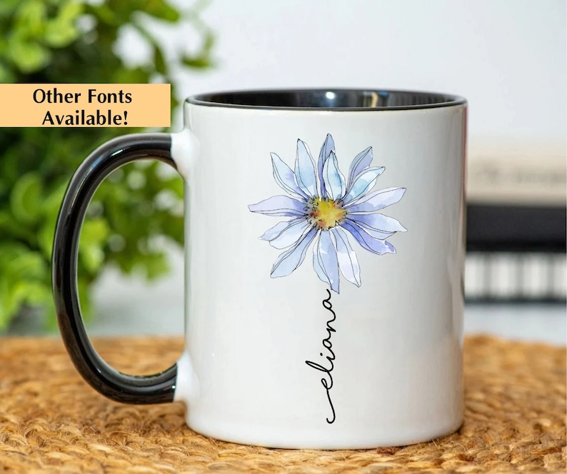 Puede incluir: Taza de cer&aacute;mica blanca con asa y borde negros. La taza presenta una ilustraci&oacute;n de acuarela de una margarita azul con un centro amarillo y el nombre "eliana" escrito en cursiva debajo de la flor.