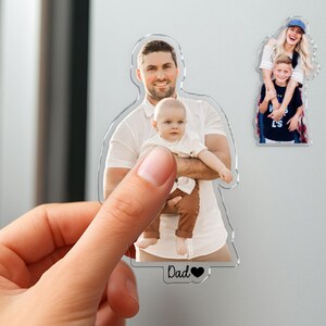 Pu&ograve; includere: Magnete fotografico in acrilico trasparente con un uomo sorridente che tiene in braccio un bambino. Il magnete ha la forma dell'uomo e del bambino. La parola "Dad" e un cuore sono in basso. Un altro magnete mostra una donna e un bambino.