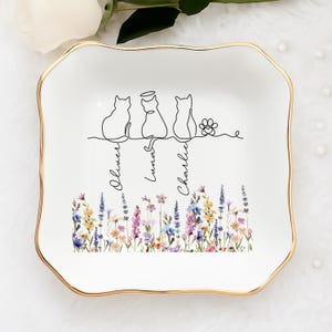 May include: White ceramic trinket dish with a gold rim. The dish features a line drawing of three cats and a paw print, with the names "Oliver", "Luna", and "Charlie" below. A floral design decorates the bottom.