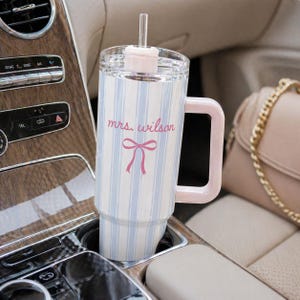 May include: A white and blue striped tumbler with a pink handle and lid, featuring the text "mrs. wilson" and a pink bow. The tumbler is in a car cup holder, next to a purse.