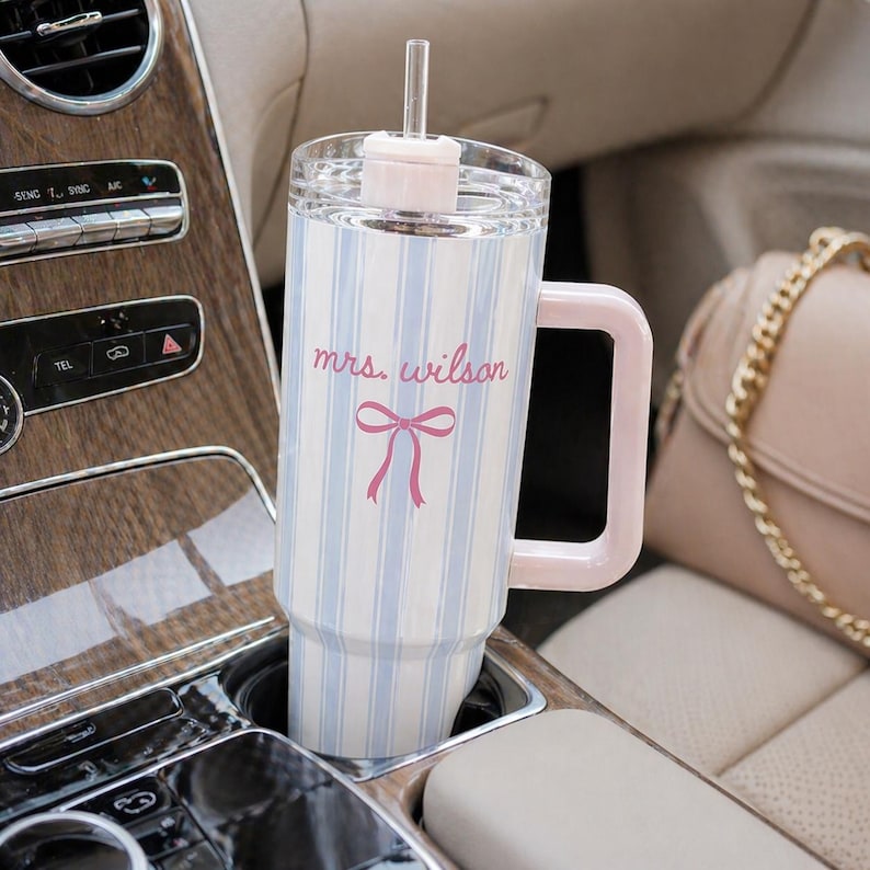 May include: A white and blue striped tumbler with a pink handle and lid, featuring the text "mrs. wilson" and a pink bow. The tumbler is in a car cup holder, next to a purse.