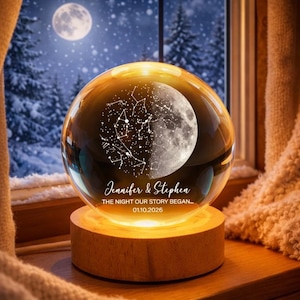 Puede incluir: Una esfera de cristal transparente con un dise&ntilde;o de luna y constelaciones, sobre una base de madera iluminada. Lleva los nombres "Jennifer & Stephen" y la fecha "01.10.2026". El fondo muestra un paisaje nevado.