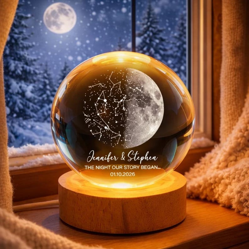 Puede incluir: Una esfera de cristal transparente con un dise&ntilde;o de luna y constelaciones, sobre una base de madera iluminada. Lleva los nombres "Jennifer & Stephen" y la fecha "01.10.2026". El fondo muestra un paisaje nevado.