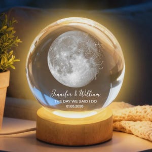 Puede incluir: Una esfera de cristal transparente con un dise&ntilde;o detallado de la luna y constelaciones, iluminada desde el interior. La esfera descansa sobre una base de madera. El texto dice "Jennifer & William THE DAY WE SAID I DO 01.05.2026".