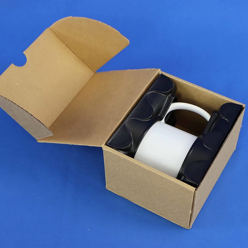 May include: A white ceramic mug inside a brown cardboard box with black protective inserts. The box is partially open, revealing the mug and its packaging. The mug is a standard size and shape.