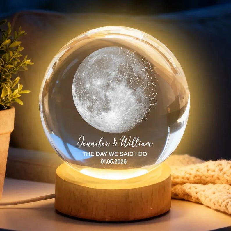 Puede incluir: Una bola de cristal transparente con un dise&ntilde;o detallado de la luna y constelaciones, iluminada por una luz c&aacute;lida. La base de madera muestra los nombres "Jennifer & William" y la fecha "01.05.2026".