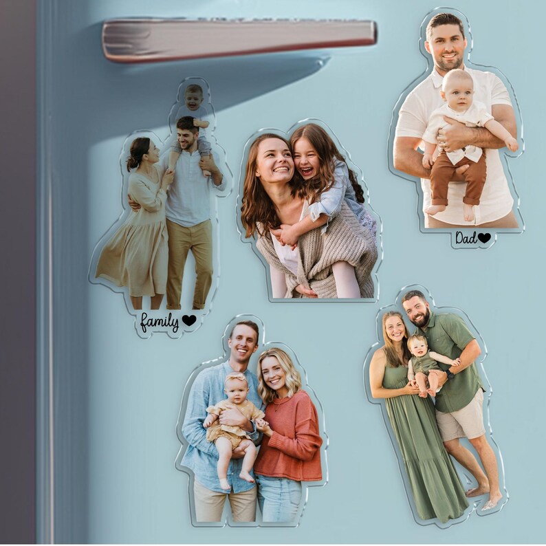 Pu&ograve; includere: Una collezione di calamite fotografiche in acrilico trasparente con ritratti di famiglia. Ogni calamita mostra una scena familiare diversa, con le parole "Family" o "Dad" e un simbolo a cuore. Le calamite sono esposte su una superficie azzurra.