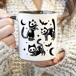 Pu&ograve; includere: Tazza in ceramica bianca con interno e manico neri. La tazza presenta un'illustrazione in bianco e nero di tre gatti scheletro con pipistrelli e lune crescenti.