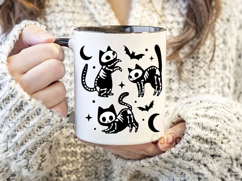 Pu&ograve; includere: Tazza in ceramica bianca con interno e manico neri. La tazza presenta un'illustrazione in bianco e nero di tre gatti scheletro con pipistrelli e lune crescenti.
