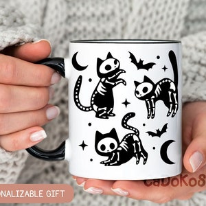 Pu&ograve; includere: Una tazza in ceramica bianca con un manico nero e illustrazioni di gatti scheletro neri. La tazza ha un bordo nero e un interno nero. Il design include stelle, lune e pipistrelli.