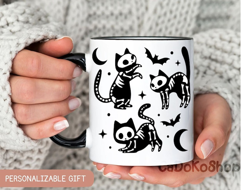 Pu&ograve; includere: Una tazza in ceramica bianca con un manico nero e illustrazioni di gatti scheletro neri. La tazza ha un bordo nero e un interno nero. Il design include stelle, lune e pipistrelli.