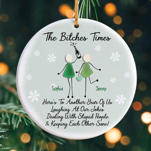 May include: A round, light green Christmas ornament with the text "The Bitches Times". It features stick figures, snowflakes, and the names "Sophia" and "Jenny". The ornament has a gold string for hanging.