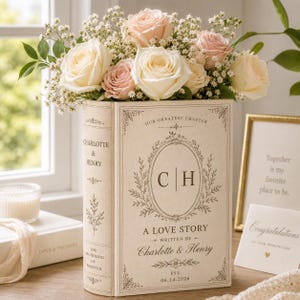 Puede incluir: Un florero con forma de libro de color crema con el texto Our Greatest Chapter, C H, A Love Story, Written By Charlotte & Henry, y Est. 14.06.2026, lleno de un ramo de rosas de color rosa p&aacute;lido y blanco y peque&ntilde;as flores de paniculata.