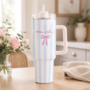 May include: A white and blue striped tumbler with a pink handle and lid. The tumbler has the text "mrs. wilson" and a pink bow graphic. A clear straw is inserted into the lid. The tumbler is on a wooden table.