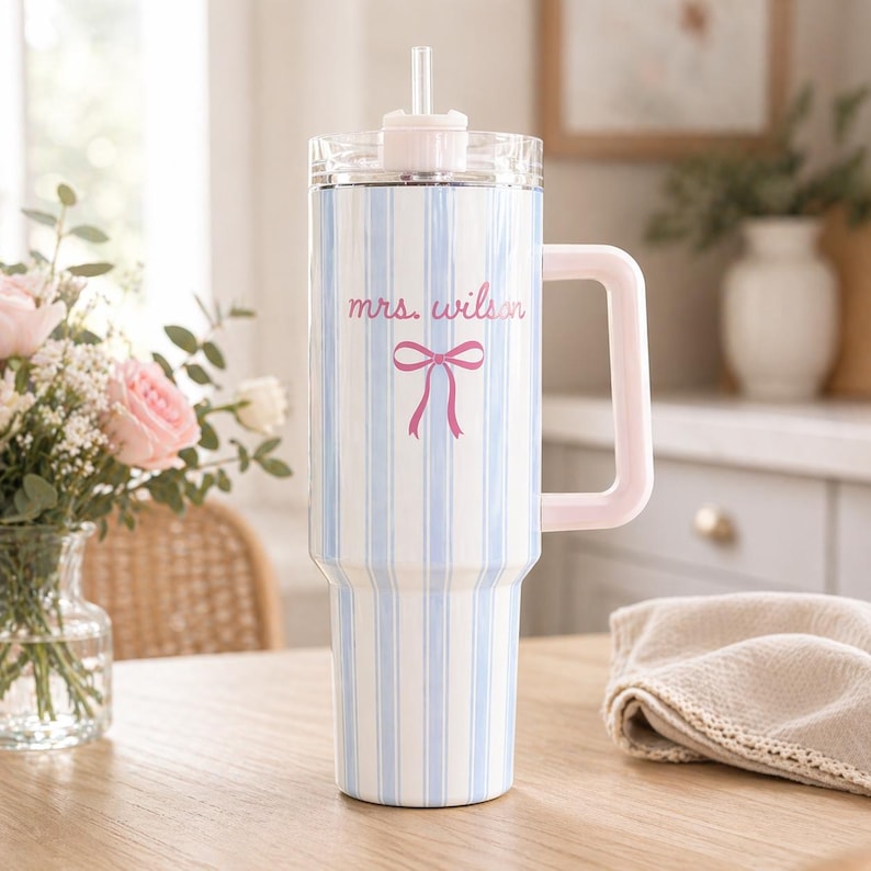 May include: A white and blue striped tumbler with a pink handle and lid. The tumbler has the text "mrs. wilson" and a pink bow graphic. A clear straw is inserted into the lid. The tumbler is on a wooden table.