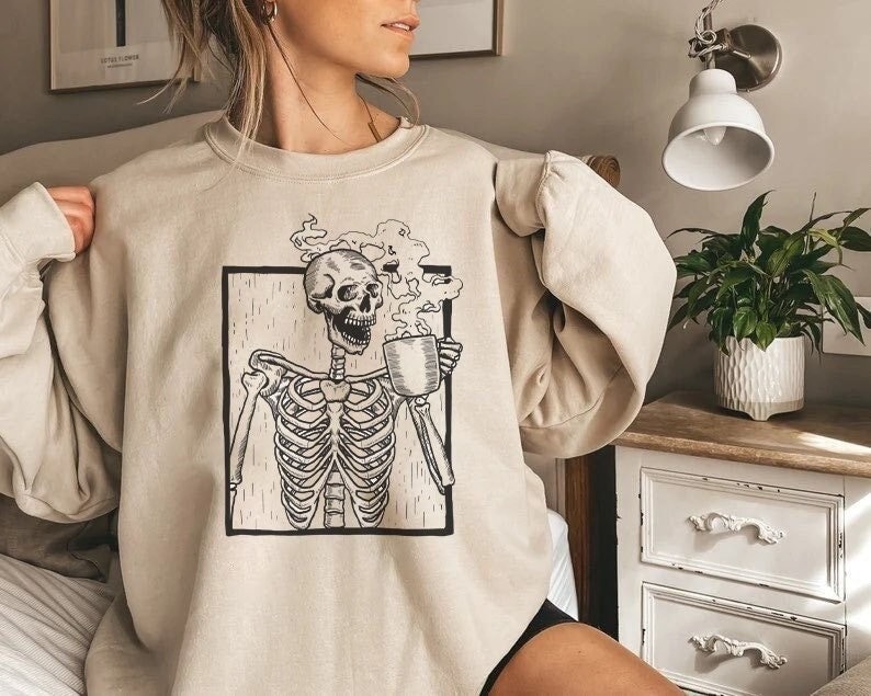 May include: A beige sweatshirt with a graphic of a skeleton holding a steaming mug. The skeleton is wearing a skull and has a ribcage. The graphic is in a black square with a white outline.