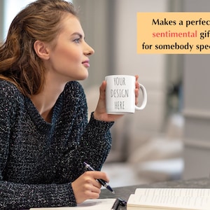 May include: A white ceramic mug with the text "YOUR DESIGN HERE" in black lettering. The mug is being held by a person wearing a black sweater.