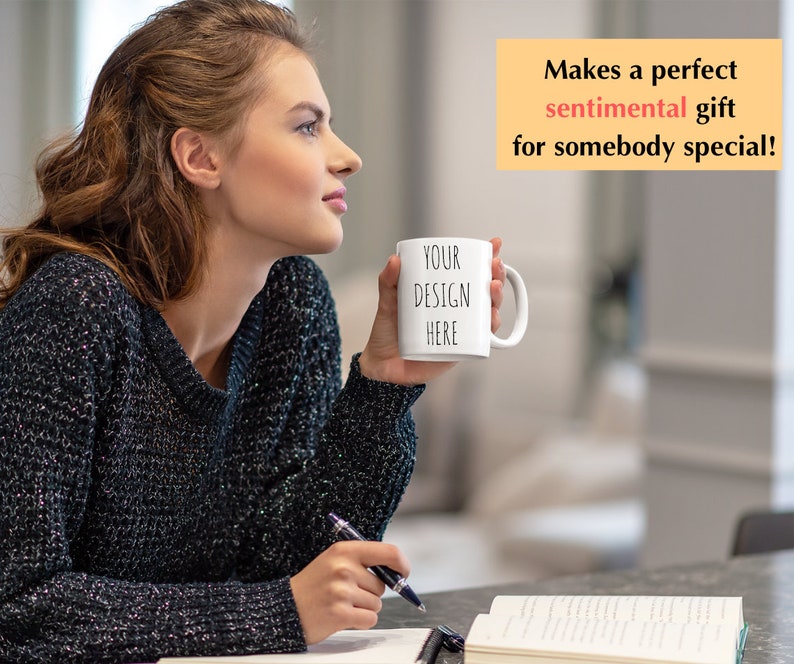 May include: A white ceramic mug with the text "YOUR DESIGN HERE" in black lettering. The mug is being held by a person wearing a black sweater.