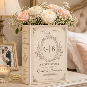 Puede incluir: Un jarr&oacute;n rectangular con forma de libro de color crema lleno de rosas de color rosa claro y blanco y paniculata. La parte frontal presenta el texto "OUR GREATEST CHAPTER", "G | B", "A LOVE STORY", "Grace & Benjamin" y "EST. 05.18.2026".