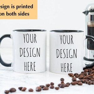 May include: Two white ceramic mugs with black rims and handles. The mugs have the text "YOUR DESIGN HERE" printed on them. The mugs are sitting on a white surface with coffee beans scattered around them.