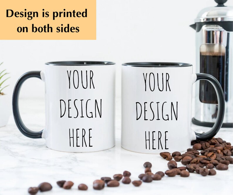 May include: Two white ceramic mugs with black rims and handles. The mugs have the text "YOUR DESIGN HERE" printed on them. The mugs are sitting on a white surface with coffee beans scattered around them.
