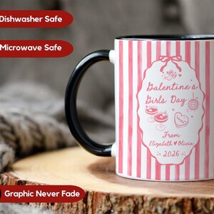 May include: A white ceramic mug with a black handle and rim. The mug features a pink and white striped pattern and a central design with the text "Valentine's Girls Day" and the names "Elizabeth & Olivia 2026".