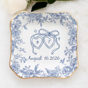 Pode incluir: Prato quadrado de cerâmica branca com design floral azul e borda dourada. O prato apresenta dois medalhões em forma de coração com as letras "B" e "T" e a data "16 de agosto de 2026". Pássaros e flores decorativas circundam o prato.