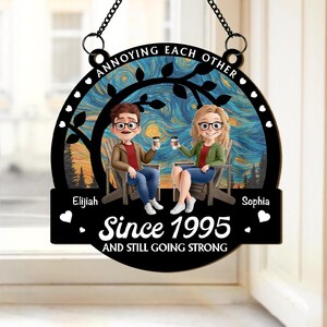 May include: A black wooden sign with the text "Annoying Each Other" and "Since 1995 AND STILL GOING STRONG." It features a couple sitting in chairs, holding coffee cups, with a starry night background. The names "Elijah" and "Sophia" are also displayed.
