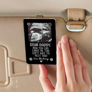 Könnte beinhalten: Ein Schwarzweiß-Ultraschallbild eines Babys in einem durchsichtigen Plastikrahmen mit dem Text "DEAR DADDY. NOW YOU CAN CARRY ME TOO CAN'T WAIT TO MEET YOU! Love, the Bump" auf schwarzem Hintergrund.