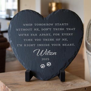 May include: A heart-shaped slate plaque with a black finish, resting on a wooden surface. The plaque features engraved text, including the name "Wilson" and the year "2025", along with paw prints. The inscription is a memorial message.