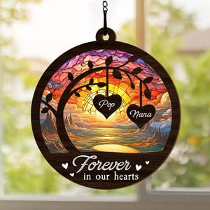 May include: A wooden suncatcher with a stained glass design of a sunset over a body of water. The suncatcher features two hearts with the words "Pop" and "Nana" inscribed on them. The text "Forever in our hearts" is also included on the suncatcher.