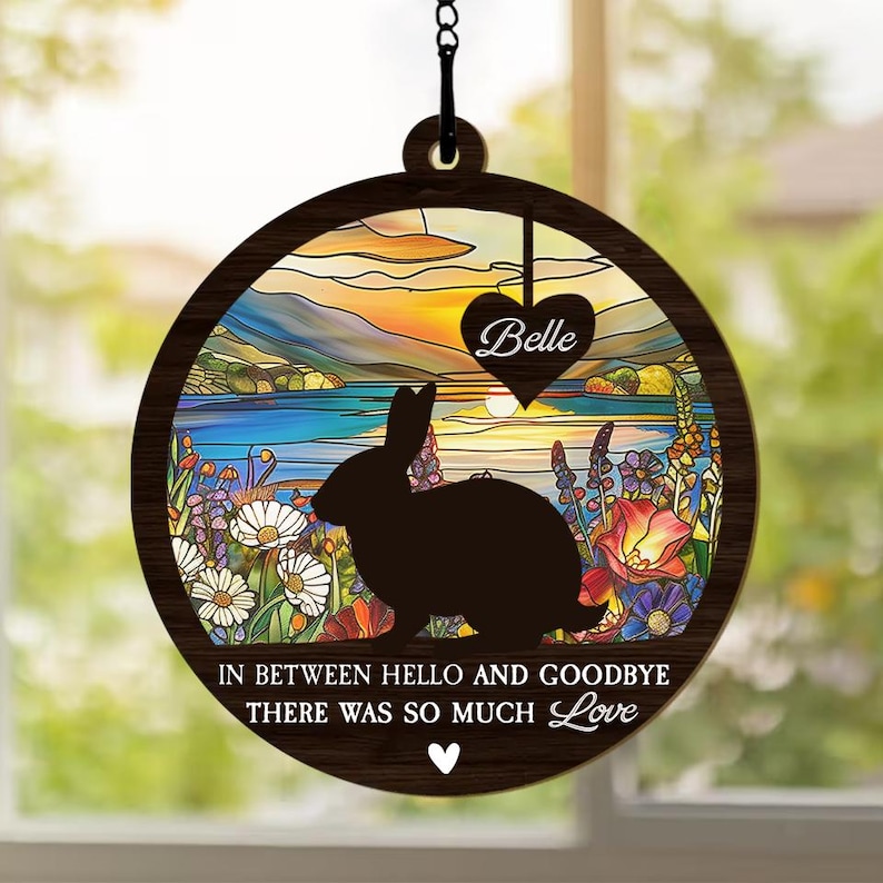 以下が含まれることがあります： 湖の上に沈む夕日を背景に、ウサギのシルエットが描かれたステンドグラスのデザインが施された木製の円形。画像の下には「In between hello and goodbye there was so much love」というテキストが印刷されています。テキストの下にはハートが印刷されています。ステンドグラスのデザインのハート型には「Belle」という文字が印刷されています。