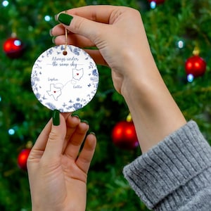 May include: A round, white ceramic Christmas ornament with a gold string, held against a green Christmas tree background. The ornament features blue floral and butterfly designs, with the text "Always under the same sky." It also includes two state outlines and names.