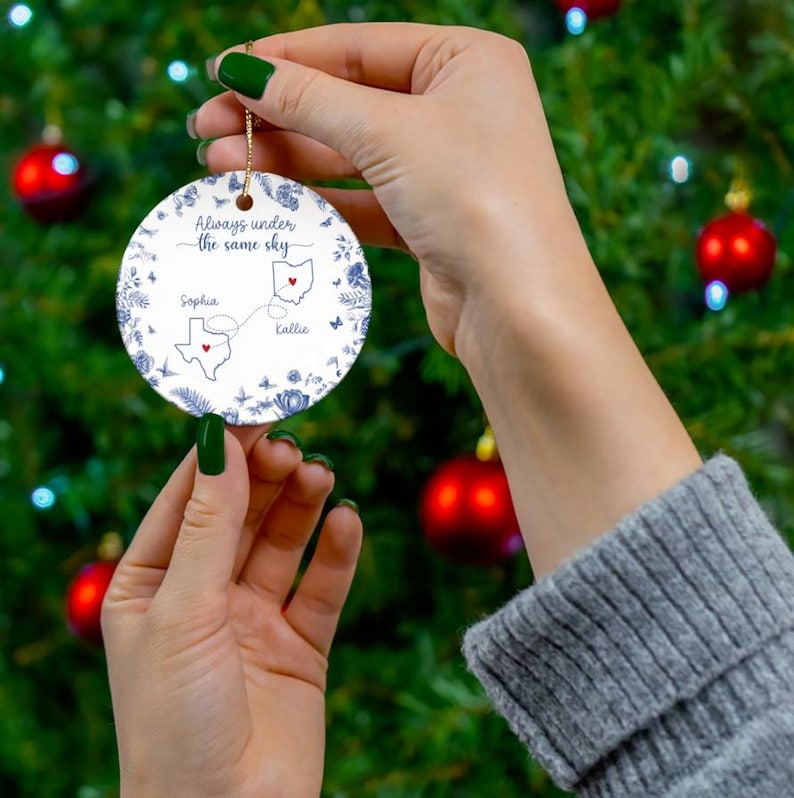 May include: A round, white ceramic Christmas ornament with a gold string, held against a green Christmas tree background. The ornament features blue floral and butterfly designs, with the text "Always under the same sky." It also includes two state outlines and names.