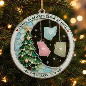 May include: A circular Christmas ornament featuring a stained-glass style tree, three state-shaped pendants with hearts, and the text "FAMILY IS ALWAYS CLOSE AT HEART" and "THE MILLERS 2026". The ornament has a silver frame and a gold hanging string.