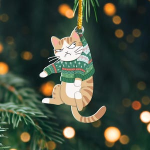 以下が含まれることがあります： 不機嫌そうな表情をした緑と赤のセーターを着た猫の漫画が描かれたクリスマスオーナメント。猫は金色の紐で吊るされています。