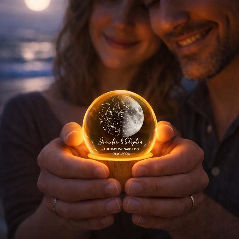 Puede incluir: Una esfera de cristal brillante con un dise&ntilde;o celestial, que presenta una media luna y constelaciones. La esfera est&aacute; iluminada desde dentro, sobre una base de madera. El texto dice "Jennifer & Stephen - THE DAY WE SAID I DO 01.10.2026".