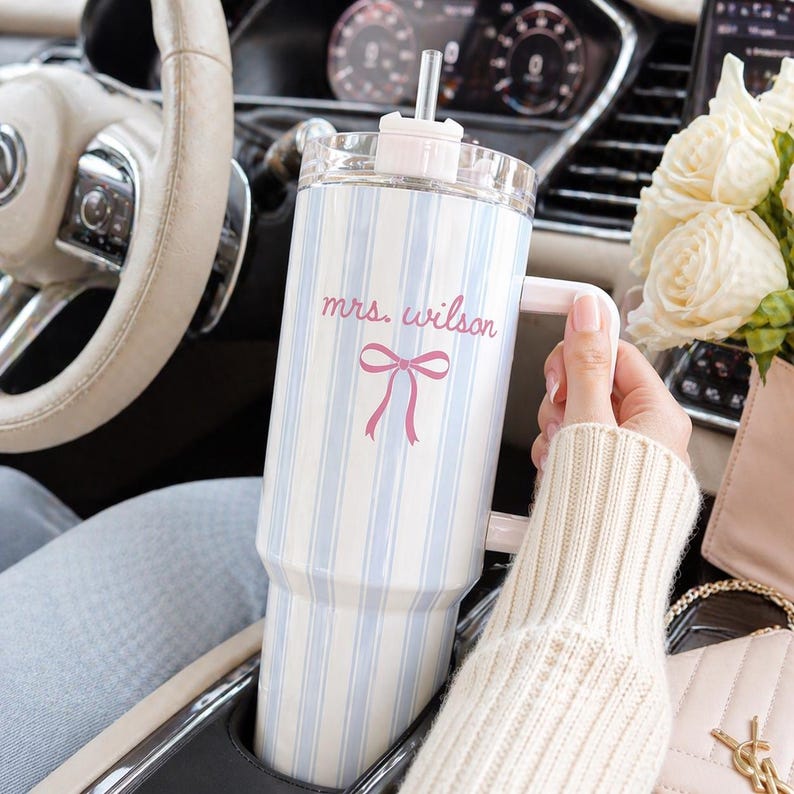 May include: A large, clear tumbler with a light blue and white striped pattern. The tumbler has a pink handle, lid, and straw. The text "mrs. wilson" and a pink bow are printed on the front.